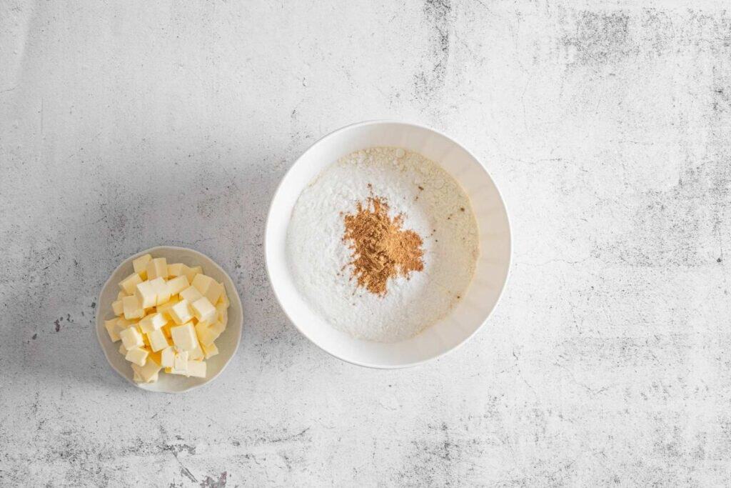 A bowl of flour topped with spices next to a small bowl of cubed butter on a light textured surface.