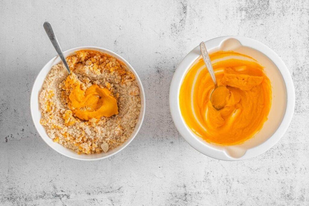 Two bowls on a white surface; the left bowl contains orange mixture with crumbled ingredients, the right bowl has a smooth orange mixture. Both have metal spoons.