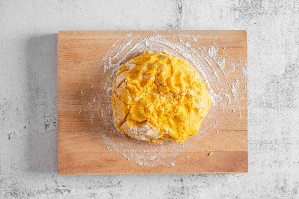 A ball of yellow dough sits on a floured wooden cutting board against a light gray textured background.