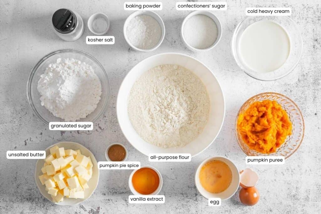 Top-down view of labeled baking ingredients, including flour, sugars, spices, pumpkin puree, butter, egg, vanilla, heavy cream, baking powder, and kosher salt in separate bowls.