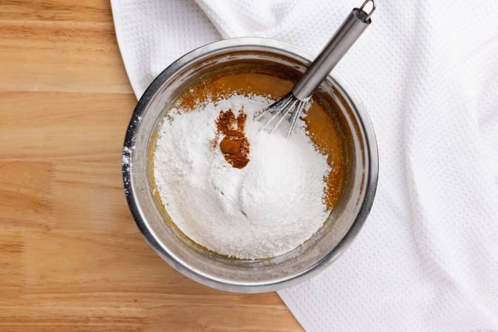 A metal bowl containing flour, cinnamon, and liquid ingredients being mixed with a whisk on a wooden surface next to a white towel.
