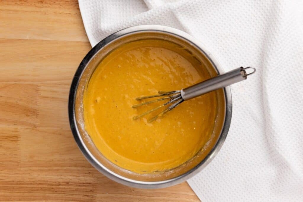 A metal bowl filled with thick yellow batter and a whisk on a wooden surface, with a white textured cloth nearby.