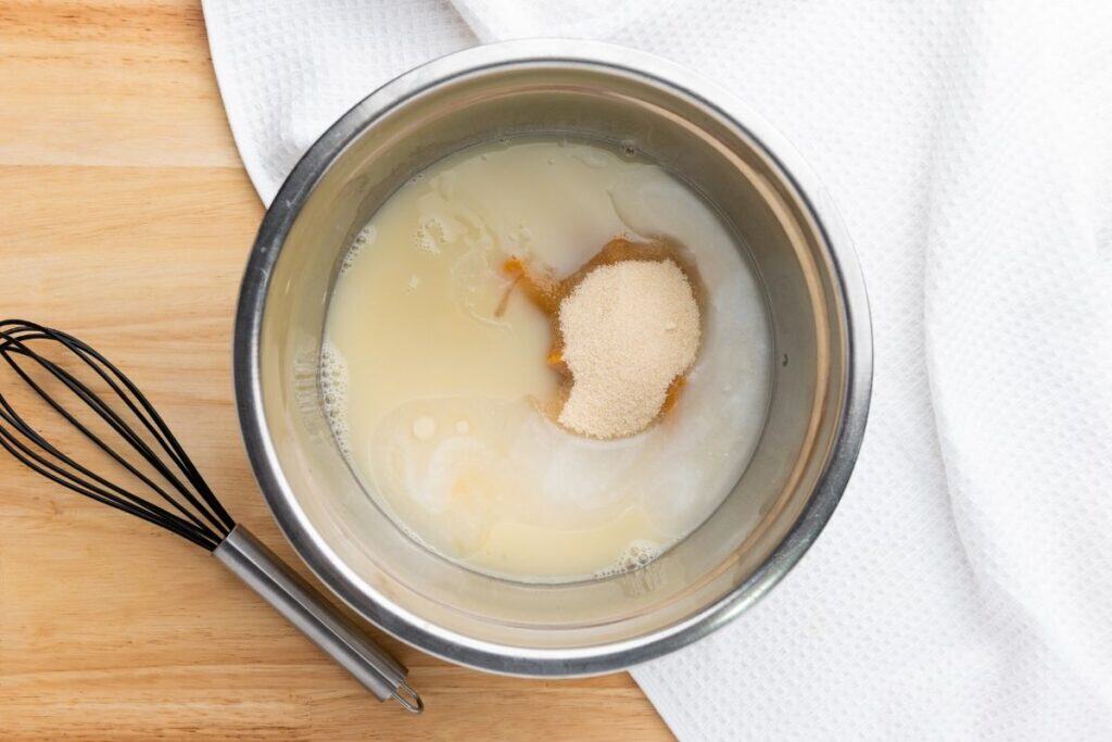 A mixing bowl contains milk, yeast, sugar, and vanilla on a wooden surface beside a whisk and a white towel.