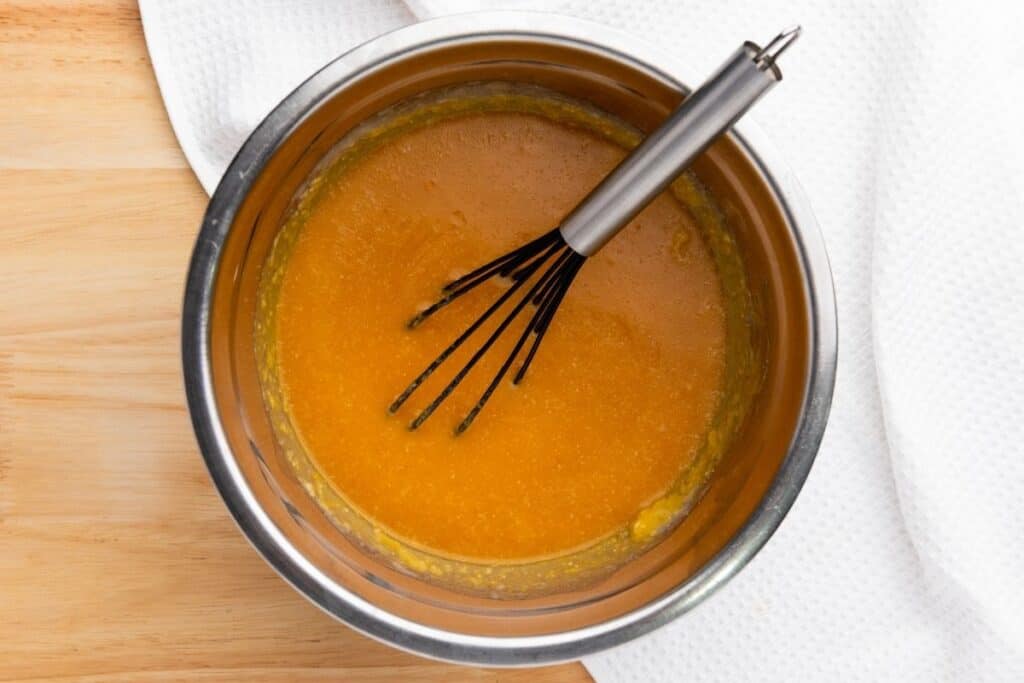 A stainless steel bowl with orange batter and a metal whisk inside, placed on a wooden surface next to a white textured cloth.