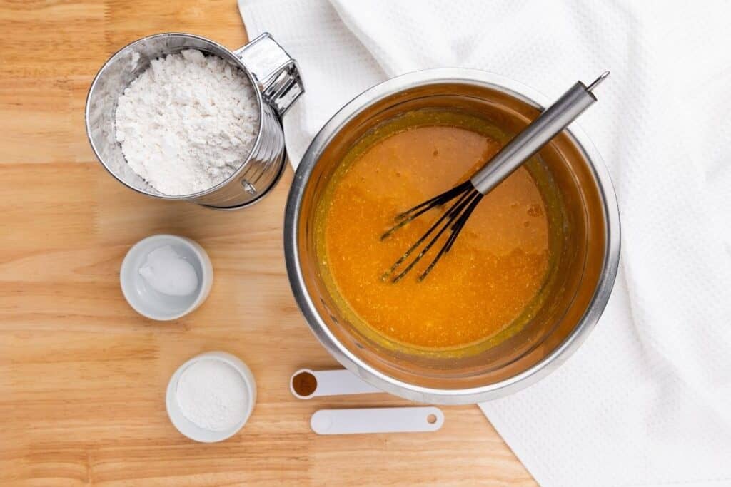 A mixing bowl with whisk and orange batter, next to a flour sifter, small bowls of baking soda and powder, and measuring spoons on a wooden surface.