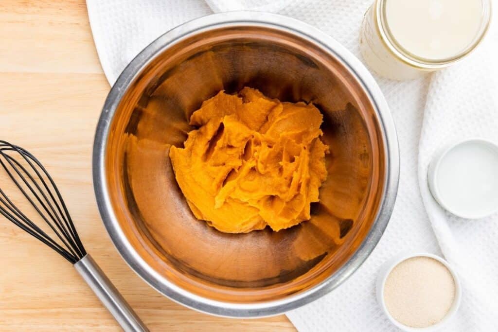 A stainless steel bowl containing smooth pumpkin puree on a wooden surface, with a whisk, a jar, a small bowl, and a white towel nearby.