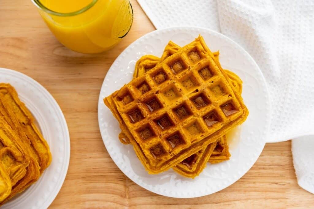 A white plate with stacked waffles sits on a wooden table next to a glass of orange juice and a white textured napkin.