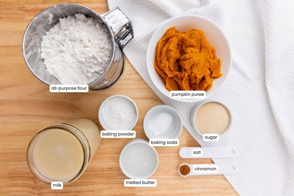 Top-down view of baking ingredients on a wooden surface, including flour, pumpkin puree, milk, baking powder, baking soda, sugar, melted butter, salt, and cinnamon, each labeled.