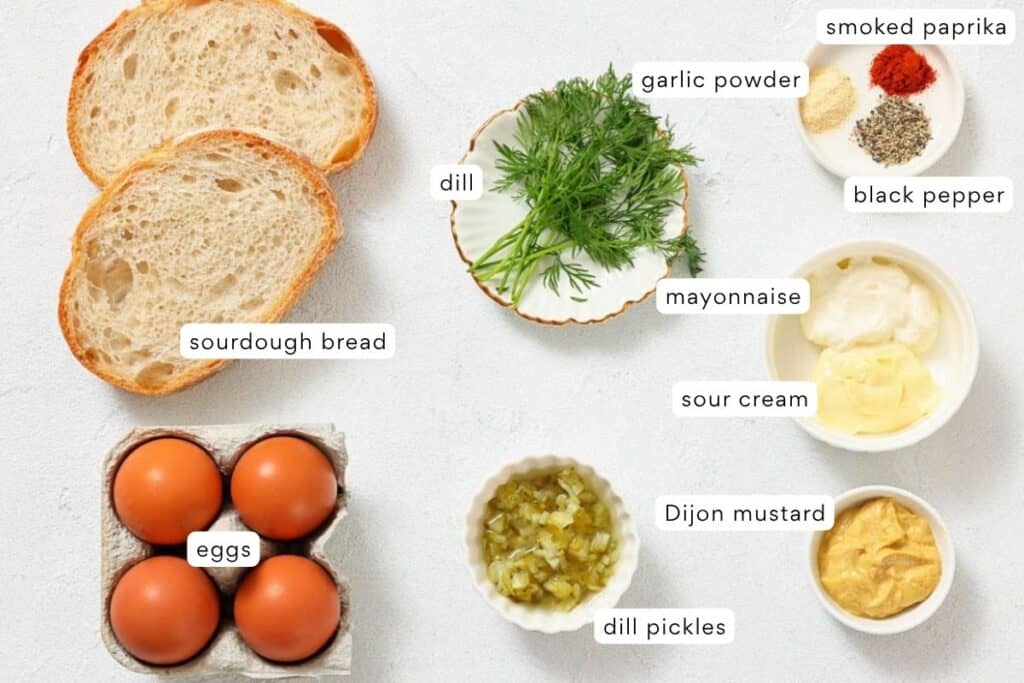 Ingredients for a recipe are laid out, including sourdough bread, eggs, dill, dill pickles, mayonnaise, sour cream, Dijon mustard, garlic powder, smoked paprika, and black pepper.