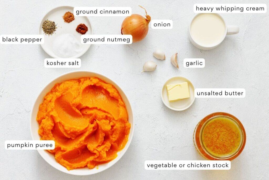Ingredients for a pumpkin soup, including pumpkin puree, cream, stock, butter, onion, garlic, black pepper, kosher salt, ground cinnamon, and ground nutmeg, arranged on a white surface.