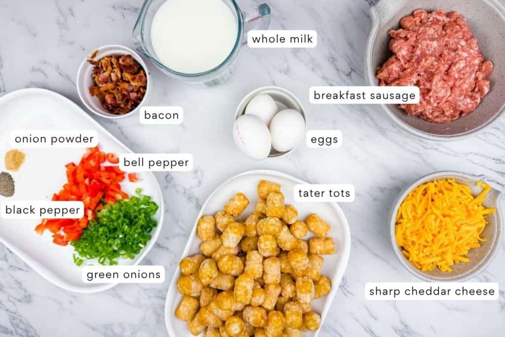 Ingredients for a breakfast casserole are arranged on a countertop, including tater tots, sausage, sharp cheddar cheese, eggs, whole milk, bacon, bell pepper, green onions, black pepper, and onion powder.