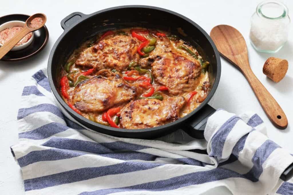 A skillet filled with cooked pork chops, sliced red and green bell peppers, and sauce, placed on a striped kitchen towel with utensils and seasonings nearby.