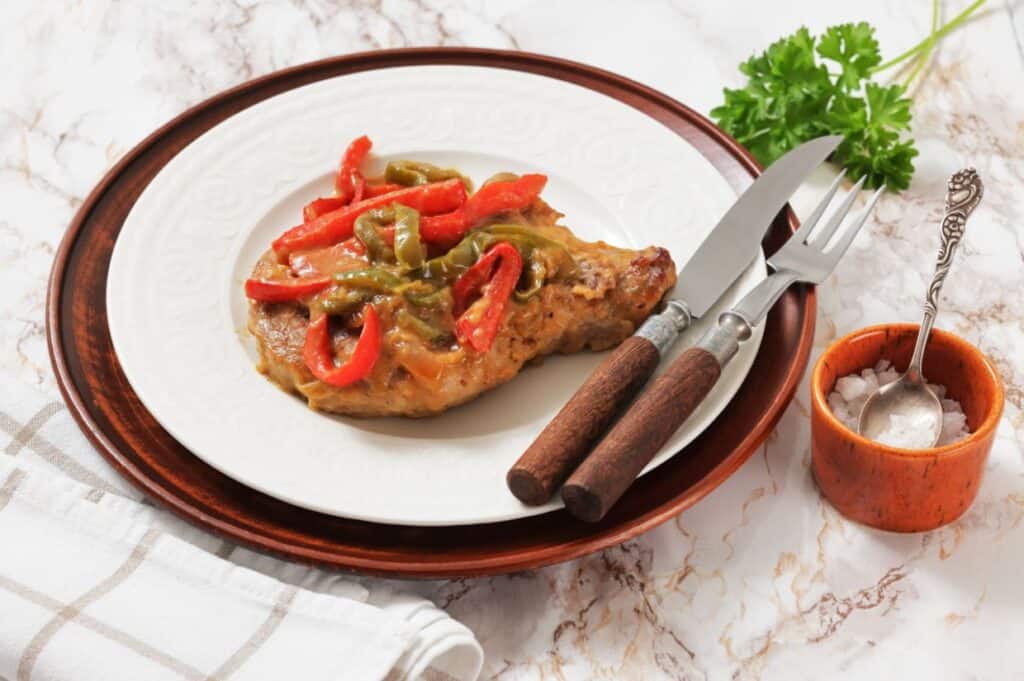 A cooked pork chop topped with red and green bell peppers on a white plate, served with a knife and fork. Salt and parsley garnish are placed beside the plate.