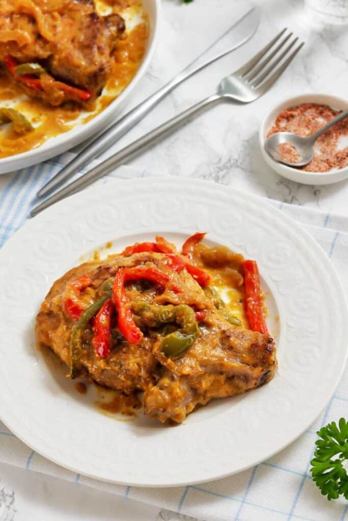 A plate with a cooked pork chop topped with red and green bell peppers and sauce, set on a white table with a fork, knife, and seasonings nearby.