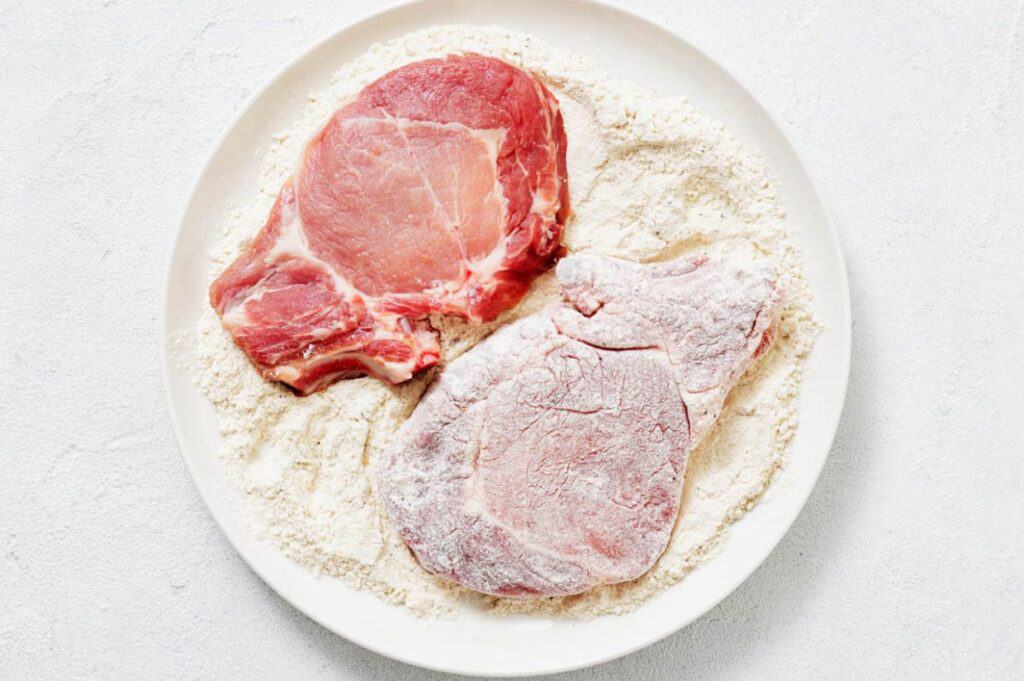 Two raw pork chops on a white plate, one fully coated with flour and the other partially coated, with excess flour on the plate.