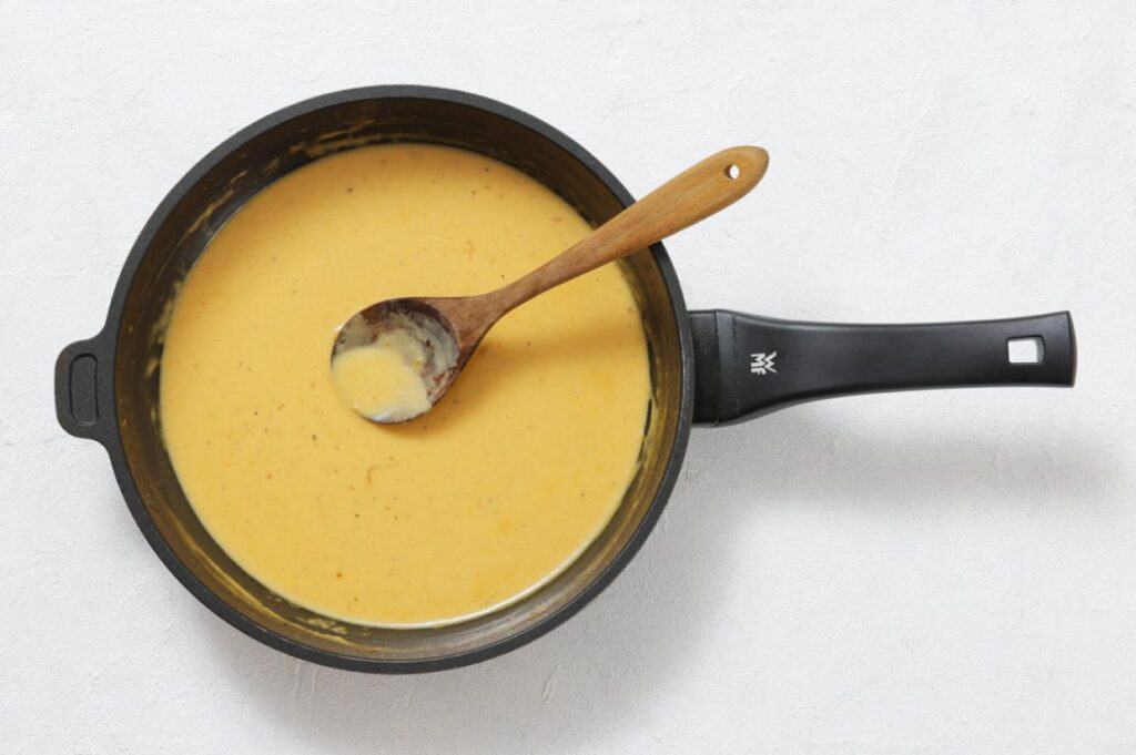 A saucepan filled with creamy yellow cheese sauce and a wooden spoon resting inside, on a white surface.