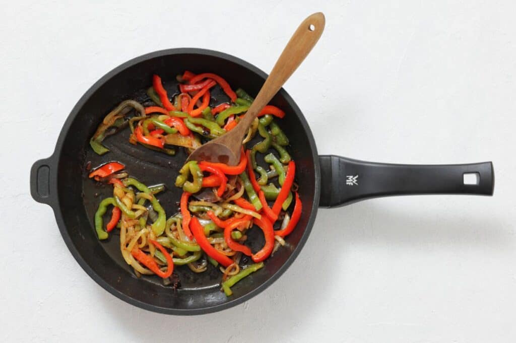 A black frying pan with cooked red and green bell pepper strips and onions, stirred with a wooden spoon, on a white background.