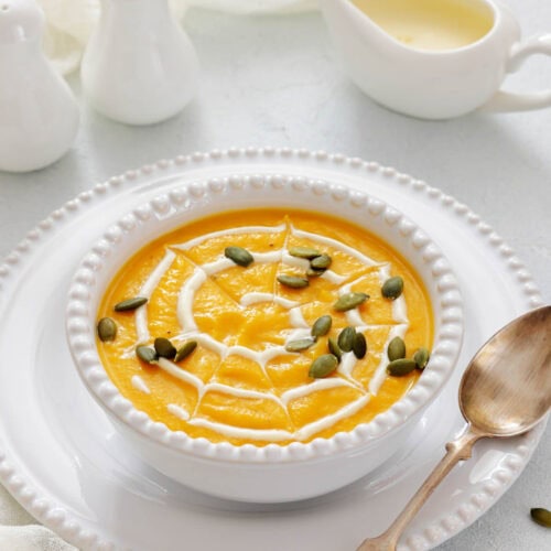 A bowl of pumpkin soup garnished with cream and pumpkin seeds, placed on a white plate with a spoon beside it; salt and pepper shakers and a sauce boat are in the background.