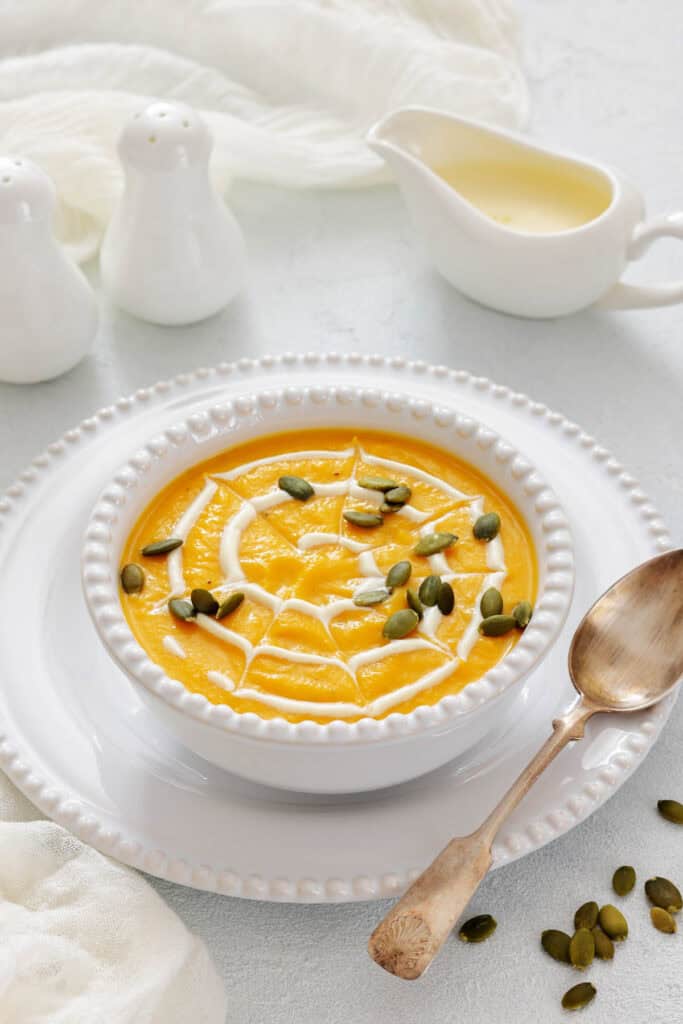 A bowl of pumpkin soup garnished with cream and pumpkin seeds, placed on a white plate with a spoon beside it; salt and pepper shakers and a sauce boat are in the background.