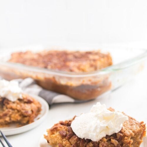 A serving of pumpkin dessert topped with whipped cream on a plate, with more dessert and a glass baking dish in the background.