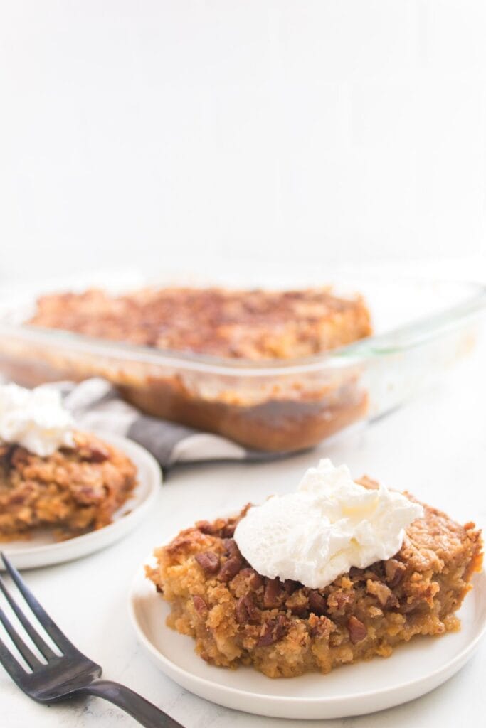 A serving of pumpkin dessert topped with whipped cream on a plate, with more dessert and a glass baking dish in the background.