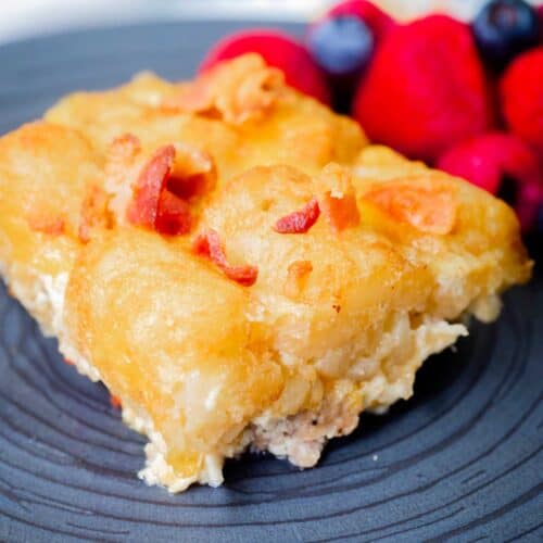 A serving of tater tot casserole with bacon bits on a black plate, accompanied by a side of mixed berries.