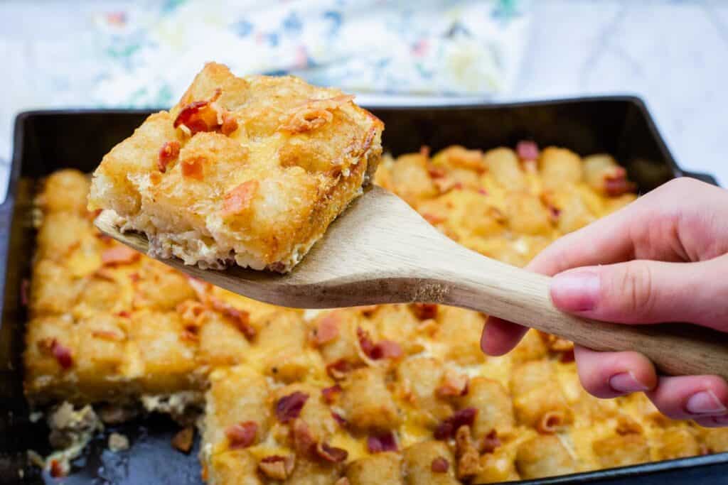 A hand holds a wooden spatula lifting a square piece of breakfast casserole topped with cheese and bacon from a baking tray.