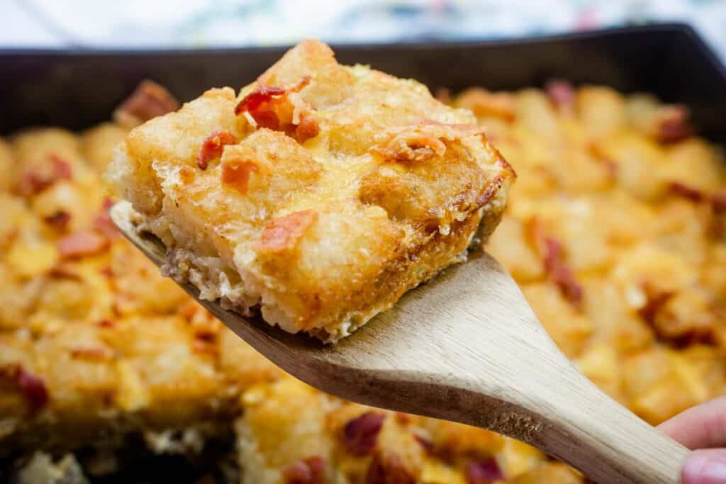 A wooden spatula holds up a square piece of baked casserole topped with crispy bits, with more casserole in the background.