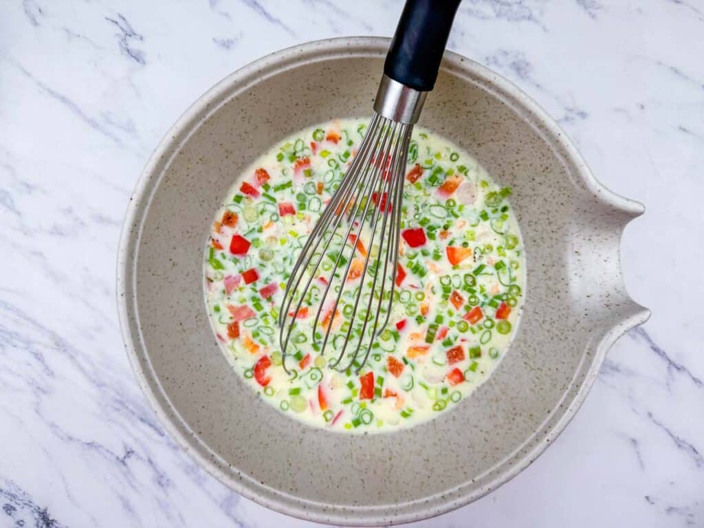 A mixing bowl with a whisk and a mixture of chopped vegetables, eggs, and milk on a white marble surface.