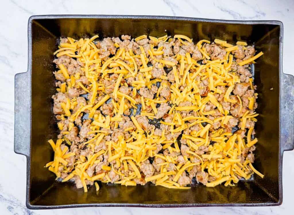A rectangular baking dish containing a layer of cooked ground meat and shredded cheddar cheese on a marble countertop.
