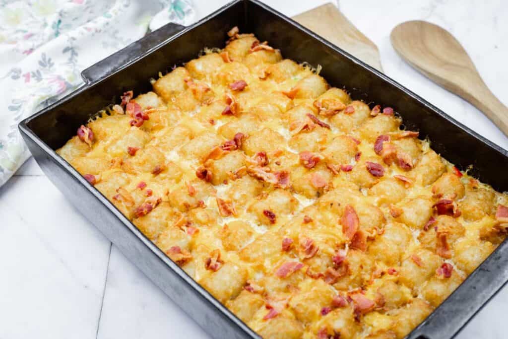 A baking dish filled with a tater tot casserole topped with melted cheese and pieces of bacon sits on a white surface next to two wooden utensils.