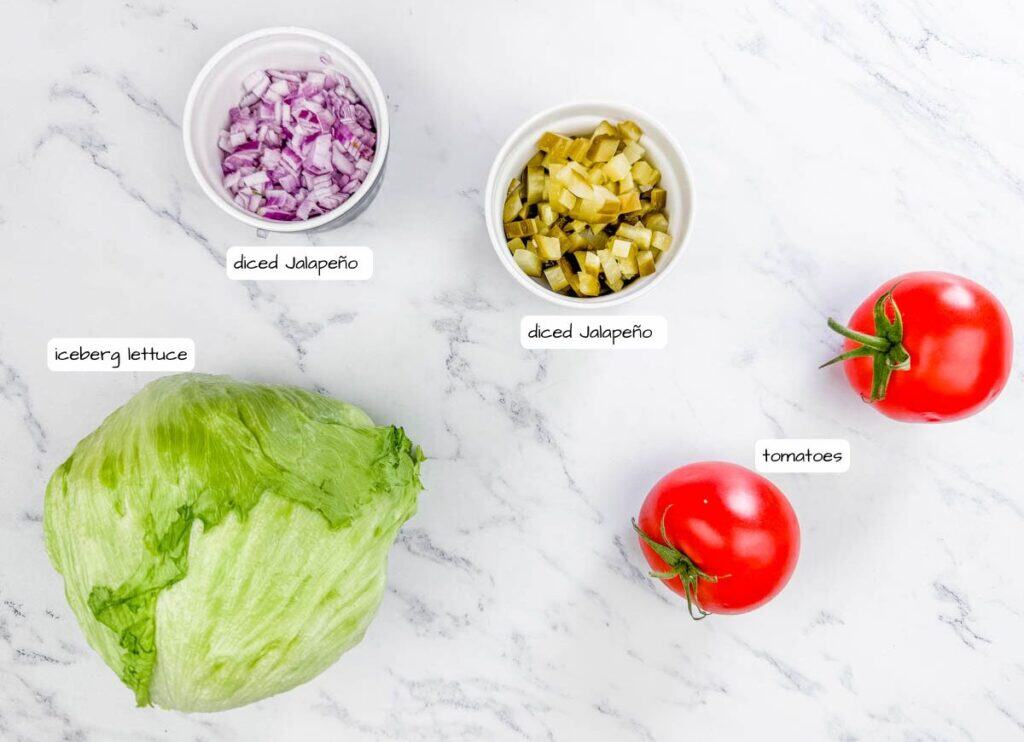 A head of iceberg lettuce, two whole tomatoes, a small bowl of diced red onion, and a small bowl of diced jalapeño on a marble surface.