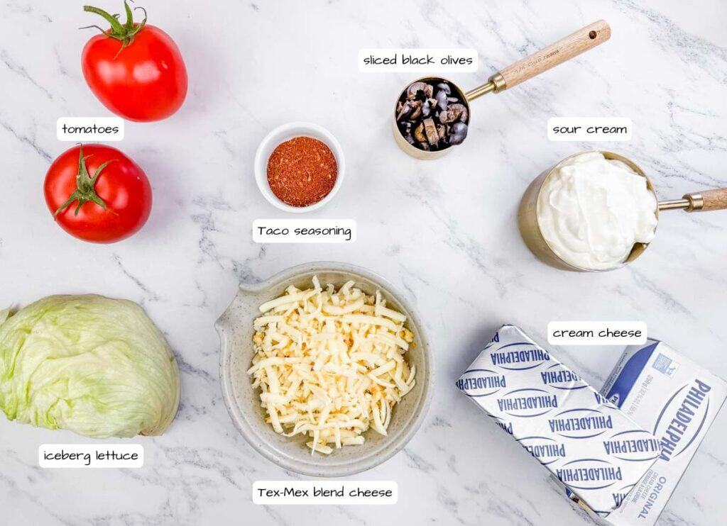 Ingredients for a Tex-Mex dish laid out on a marble surface: tomatoes, iceberg lettuce, taco seasoning, sliced black olives, sour cream, cream cheese, and shredded cheese.