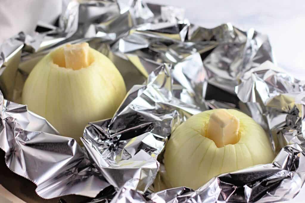 Two peeled onions with butter on top sit in crumpled aluminum foil, prepared for baking or roasting.