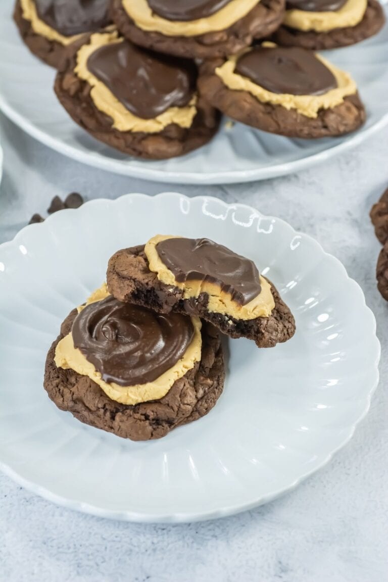 A chocolate cookie topped with a layer of peanut butter and chocolate frosting sits on a plate, with one cookie partially eaten; more cookies are visible in the background.