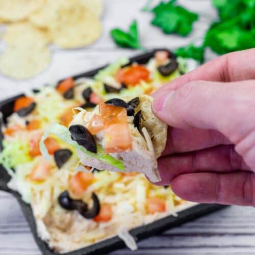 A hand holds a tortilla chip topped with layered dip including shredded cheese, olives, lettuce, and diced tomatoes. More chips and dip are in the background.
