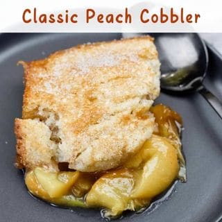 A serving of classic peach cobbler on a dark plate, featuring golden-brown, sugar-crusted biscuit topping over warm, gooey peach filling. A spoon rests beside the dessert, ready for digging in.