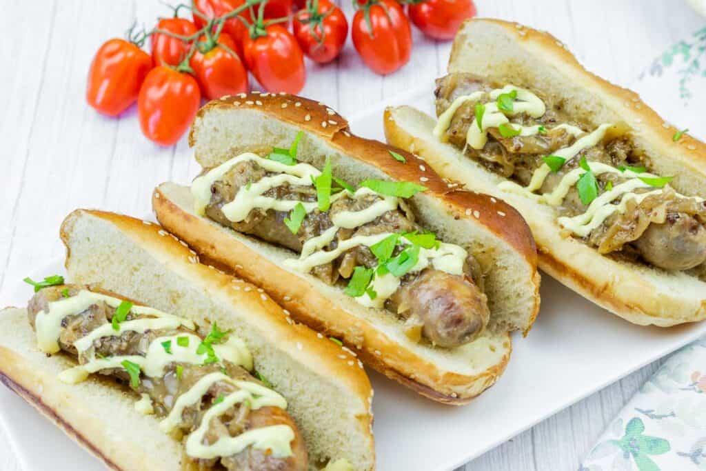 Three sausage sandwiches in toasted buns topped with grilled onions, chopped parsley, and a drizzle of sauce, served on a white plate with cherry tomatoes in the background.