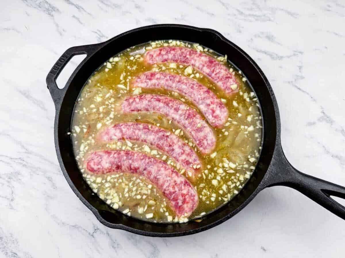 Five raw sausages are arranged in a cast iron skillet filled with chopped onions and liquid on a white marble surface.