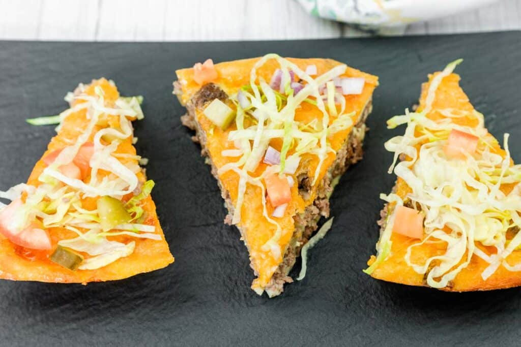 Three triangular slices of taco pizza topped with ground beef, melted cheese, shredded lettuce, diced tomatoes, onions, and pickles, arranged on a black slate surface.