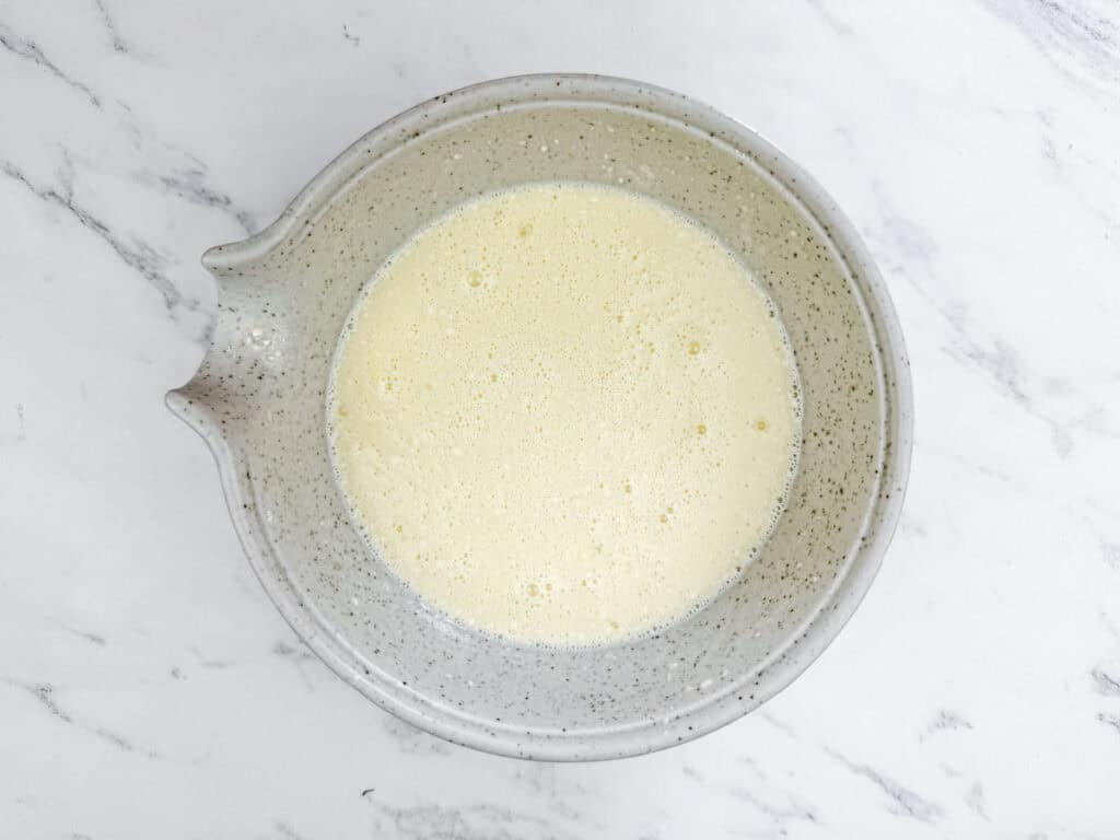 A speckled gray mixing bowl filled with a pale yellow, frothy liquid sits on a white marble surface.