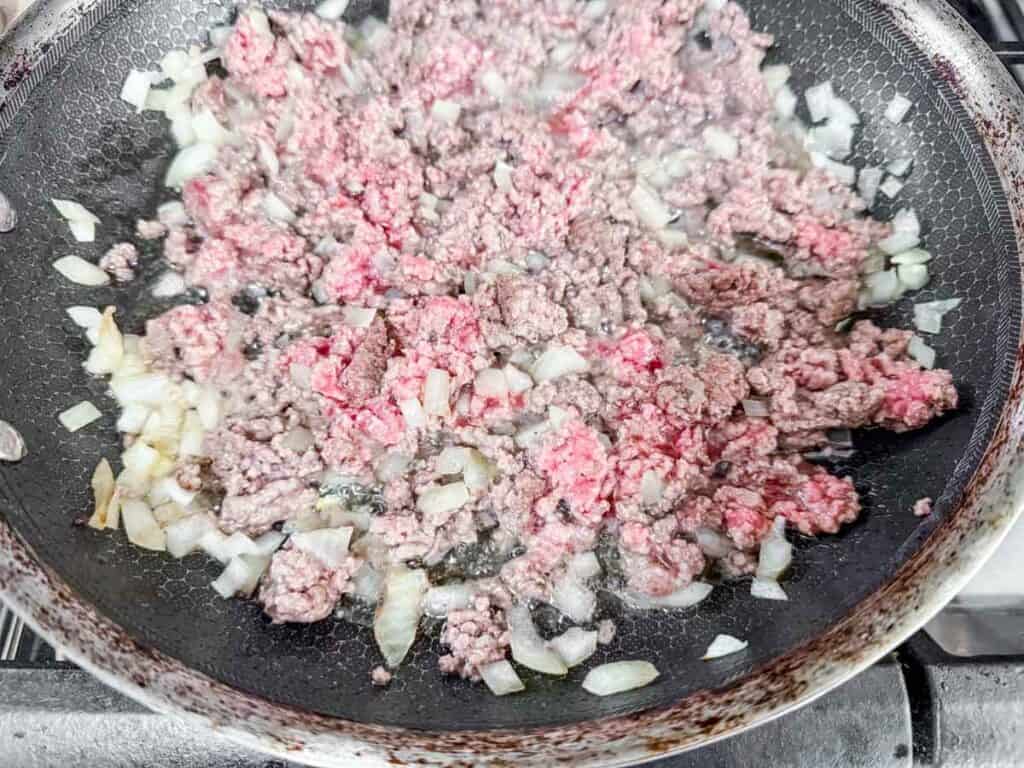 Ground beef and chopped onions being cooked in a frying pan on a stovetop.