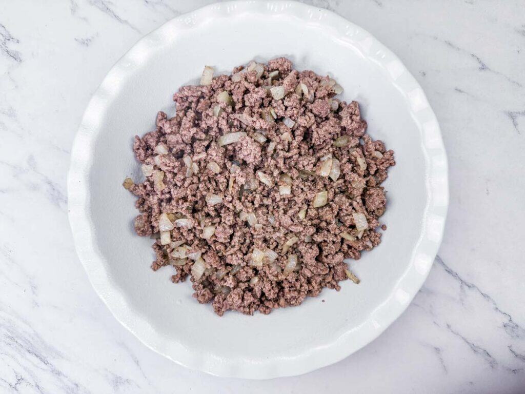 A white plate filled with cooked ground beef mixed with diced onions, placed on a marble countertop.
