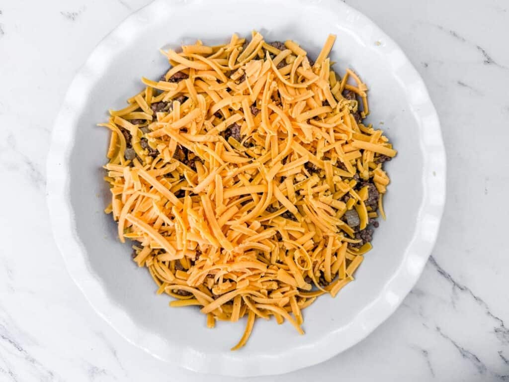 A white dish filled with shredded cheddar cheese and a mixture of ground beef on a marble surface.