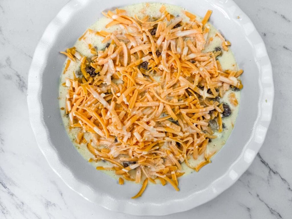 A pie dish filled with an unbaked mixture of shredded cheese, vegetables, and creamy sauce, placed on a marble countertop.