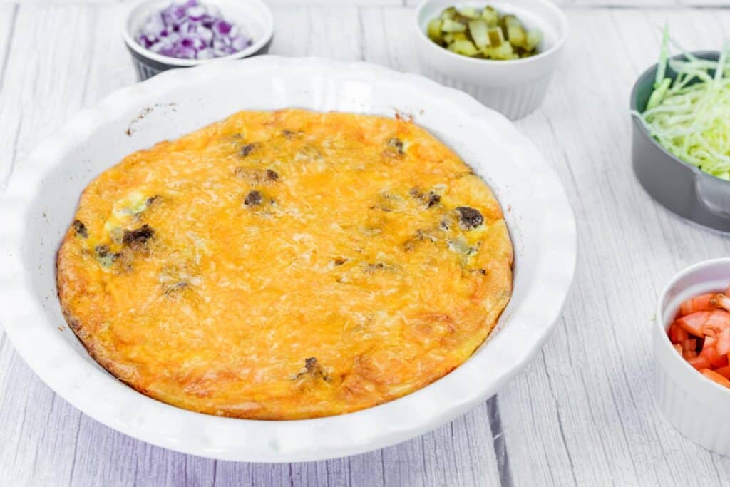 A baked casserole with a golden cheese topping in a white dish, surrounded by small bowls of chopped onions, pickles, shredded lettuce, and diced tomatoes.