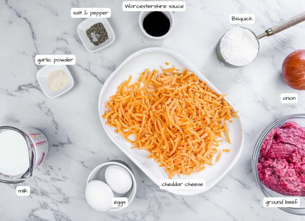 Various labeled ingredients for a recipe are arranged on a marble surface, including cheddar cheese, eggs, ground beef, milk, Bisquick, onion, garlic powder, salt and pepper, and Worcestershire sauce.