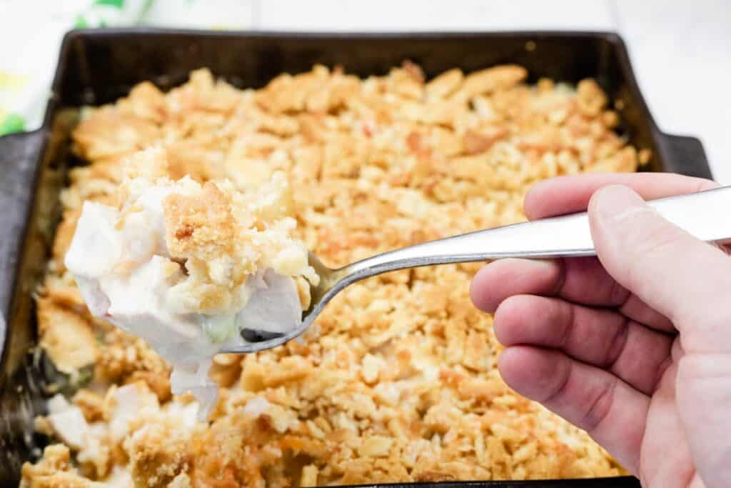 A hand holding a spoon over a casserole dish with chicken and cheese.
