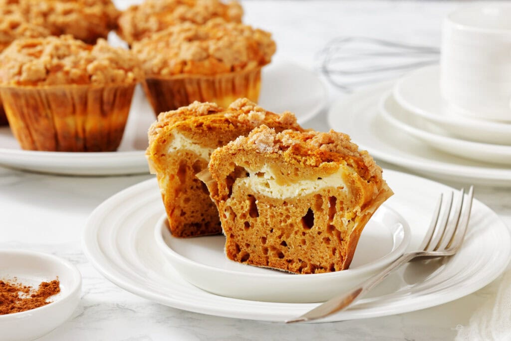 A pumpkin muffin with cream cheese filling is halved on a white plate, with a fork beside it. Whole muffins and stacked plates are in the background.
