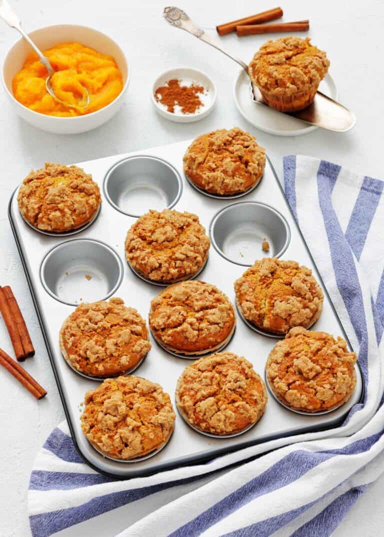 A muffin tin filled with nine streusel-topped muffins sits on a table beside a bowl of pumpkin puree, a bowl of cinnamon, a single muffin on a spatula, and a striped kitchen towel.